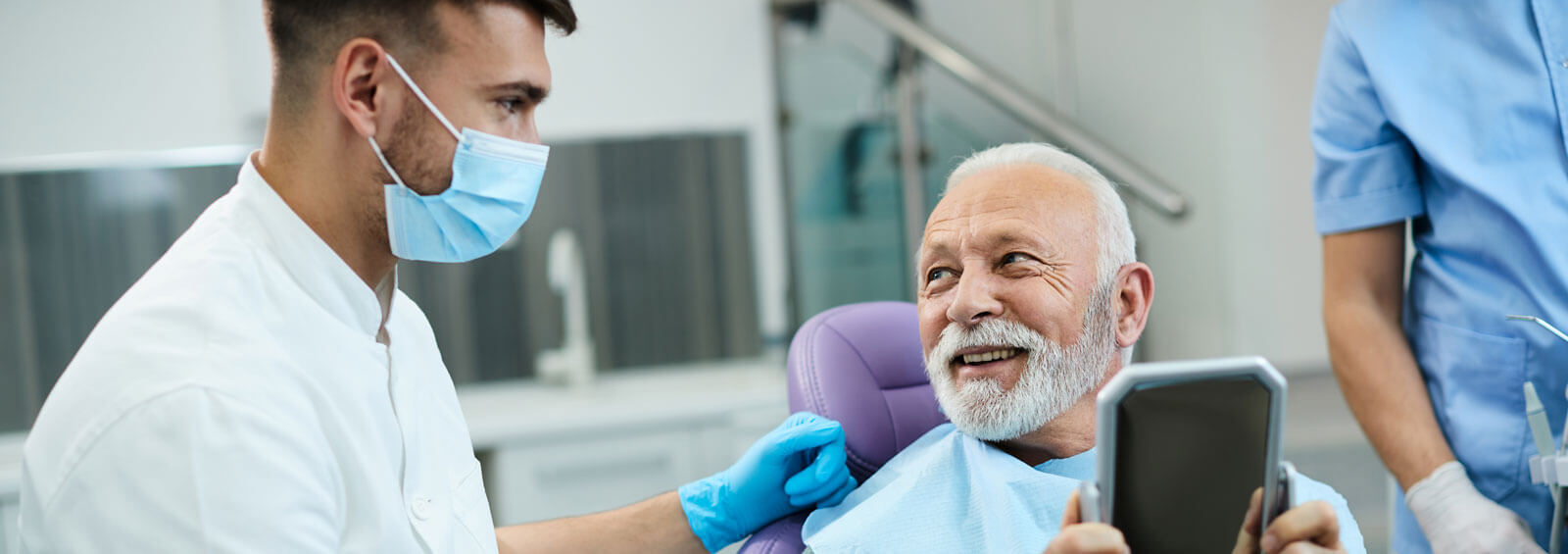 Satisfied Senior Man Talks With Dentist While Looking His Teeth In Mirror At Dentist's Office.